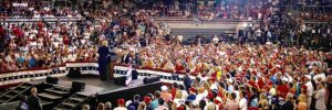 Trump stands on small stage with his back to camera, surrounded by larger audience.