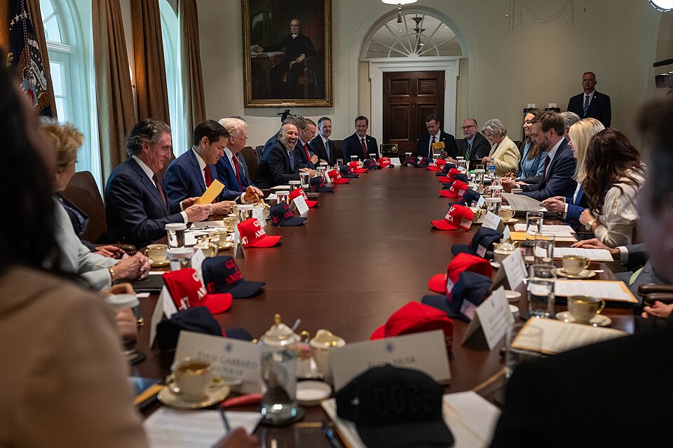 People in suits seated around a very large table, with a red baseball cap on the table in front of almost every person.