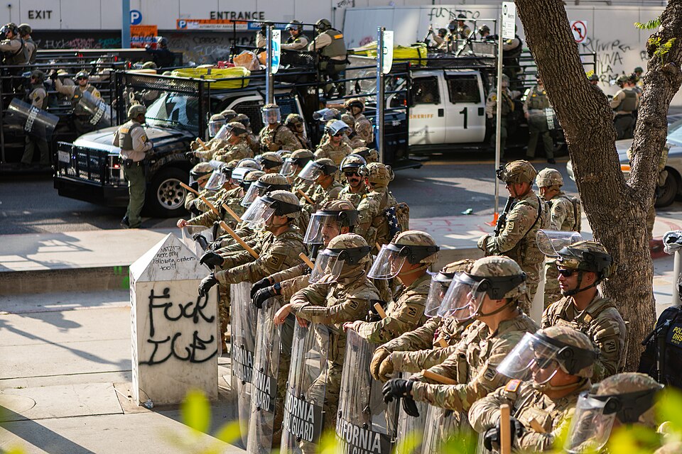 *A wall of uniformed California National Guard troops in riot gear, by pillar with "Fuck ICE" graffiti.