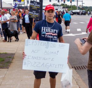 Man wearing Make America Great Again hat and t-shirt that says "Hillary for Prison 2016" and holding sign that says "(((Epstein))) did not act alone! Investigate #Pizzagate!!!"