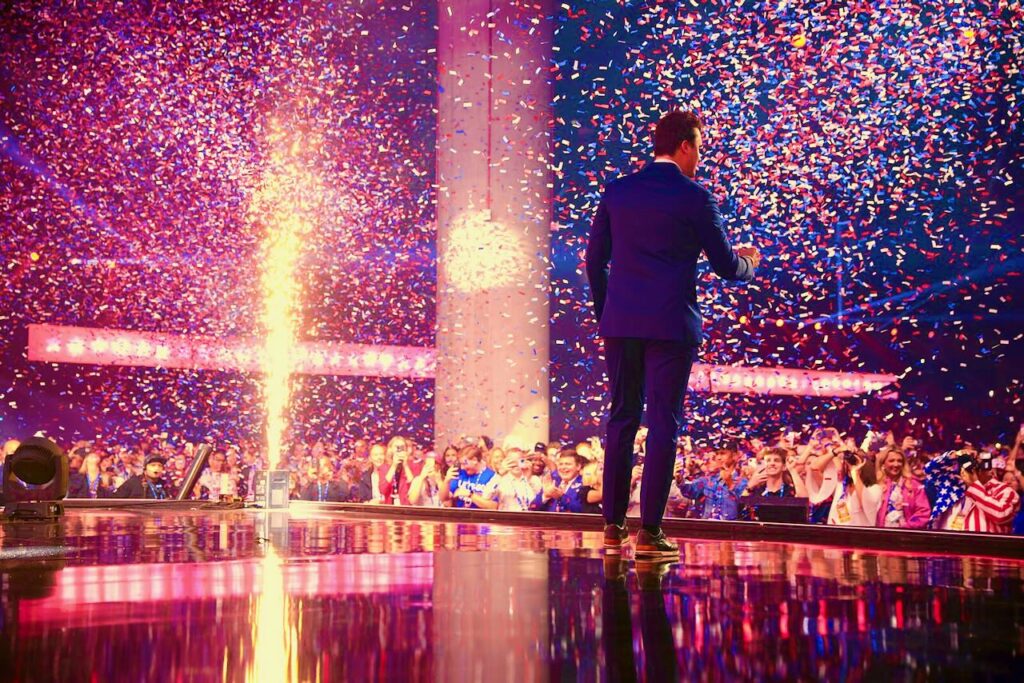 Charlie Kirk standing on stage facing audience with his back to the camera, with confetti falling everywhere