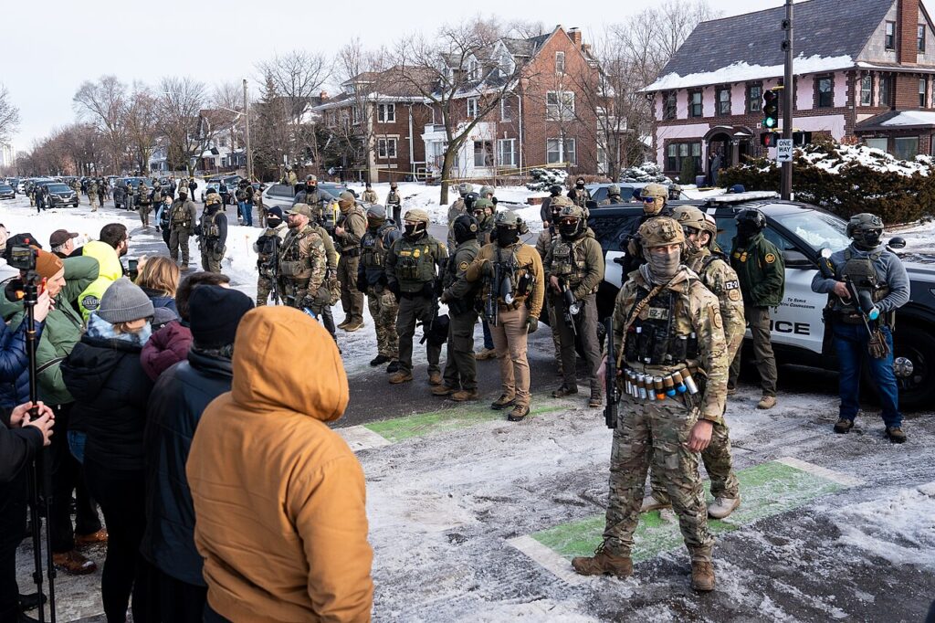 Uniformed ICE officers face protesters along snow-covered street.