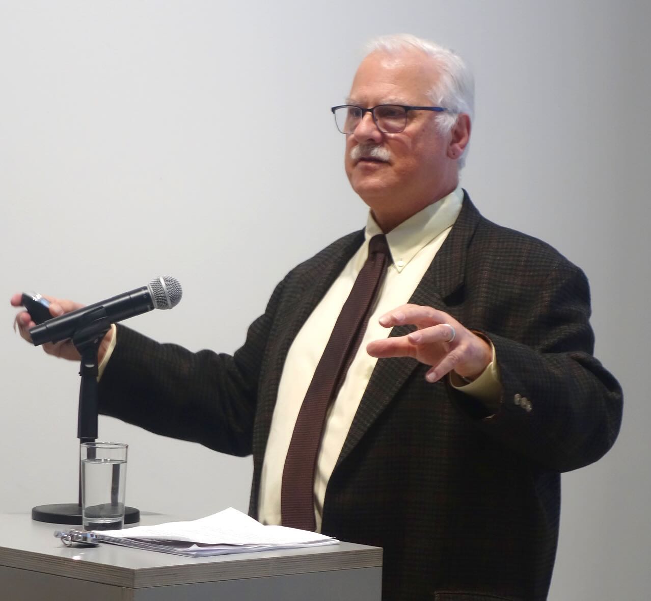 Chip Berlet wearing a jacket and tie, speaking in front of a microphone, gesturing with his hands.