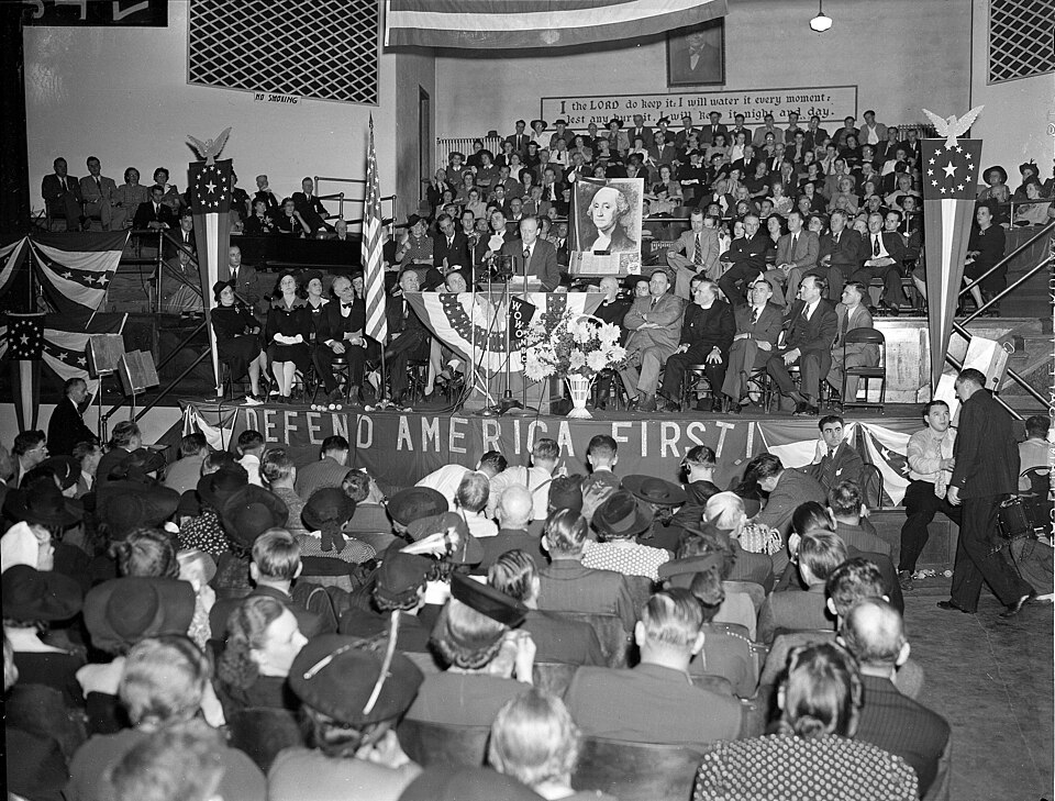 Man at podium at large indoor rally; banner in front of speaker reads "Defend America First", portrait of George Washington is behind speaker.